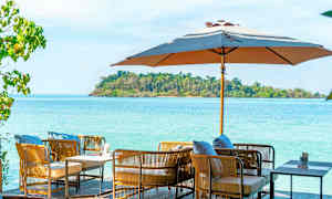 Udendørsmøbler med puder under en parasol med hav og grøn ø i baggrunden. (AI-genereret tekst)