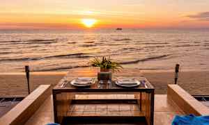 Et dækket bord med to tallerkener og bestik står ved havet under en solnedgang. (AI-genereret tekst)