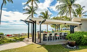 En udendørs bar ved en tropisk strand med palmer og hav i baggrunden. (AI-genereret tekst)