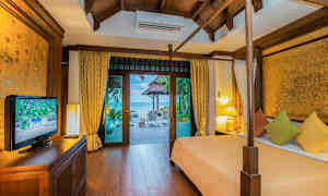 Et soveværelse med trækonstruktion, der har udsigt mod en terrasse og havet udenfor. Værelset indeholder en seng med farverige puder, et TV-skab og lamper. (AI-genereret tekst)