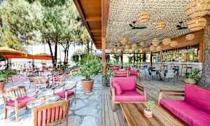 Udendørsrestaurant med træborde og stole, lyserøde puder, omgivet af træer og parasoller. (AI-genereret tekst)