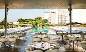 Billede af en udendørsrestaurant med borde og stole, ved en stor pool omgivet af liggestole og parasoller, med grøntområder og bygninger i baggrunden. (AI-genereret tekst)