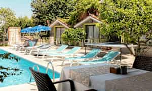 En udendørs pool ved siden af liggestole, borde og stole placeret tæt på små bygninger. (AI-genereret tekst)