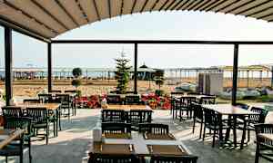 En udendørs servering med borde og stole, i baggrunden en sandstrand og havet. (AI-genereret tekst)