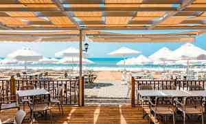 Udendørs servering med borde, stole og parasoller tæt på havet. (AI-genereret tekst)