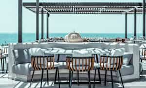 Et udendørs bord med kurvestole og bløde puder placeret på en terrasse med udsigt over havet. (AI-genereret tekst)