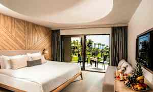 Et moderne hotelværelse med en stor dobbeltseng, en sofa og et bord med frugtfad. Værelset har skydedøre af glas, der fører ud til en balkon med udsigt over en frodig have og havet i baggrunden. (AI-genereret tekst)