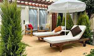 To liggestole med hvide hynder og puder står på en træterrasse under en parasol, omgivet af grønne planter. (AI-genereret tekst)