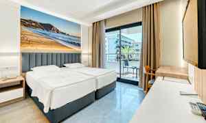 Hotelværelse med to enkeltsenge, balkon i baggrunden og stort fotografi af en strand på væggen. (AI-genereret tekst)