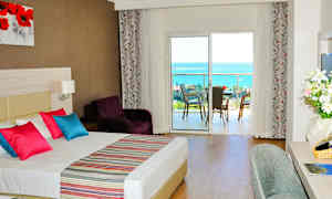 Hotelværelse med dobbeltseng, farverige puder og udsigt til en balkon med havudsigt. (AI-genereret tekst)