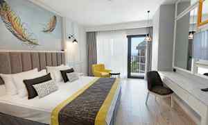 Et hotelværelse med en dobbeltseng, gul lænestol, skrivebord og spejl samt balkon med havudsigt. (AI-genereret tekst)