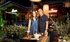 To personer ved et dækket bord med en kage og tændte lys på en restaurant med grønne omgivelser og lamper. (AI-genereret tekst)