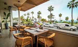Billedet viser en restaurant med udsigt over en gade med parkerede og forbipasserende biler samt palmer i baggrunden. (AI-genereret tekst)