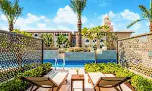 To liggestole ved en privat pool, omgivet af grønne planter og med udsigt til en bygning med middelhavsinspirerede arkitekturelementer. (AI-genereret tekst)