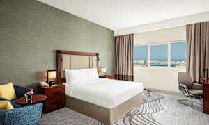 Et hotelværelse med en dobbeltseng, natbord, lænestol og vindue med udsigt over en by. (AI-genereret tekst)