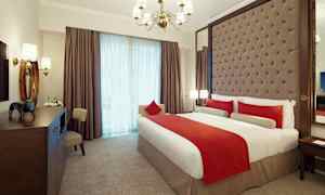 Et elegant indrettet hotelværelse med en stor seng redt med hvide og røde sengesæt. Værelset har en polstret sengegavl, en lysekrone og store vinduer med gardiner. (AI-genereret tekst)