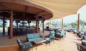 En udendørs bar med siddegrupper og trætak, omgivet af palmer og byens skyline i baggrunden. (AI-genereret tekst)