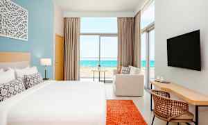 Et moderne hotelværelse med en stor seng, en sofa, skrivebord og et TV. Udsigten fra rummet viser en strand og havet. (AI-genereret tekst)