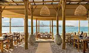 En strandrestaurant med træborde og stole, udsigt over havet og en by i baggrunden. (AI-genereret tekst)