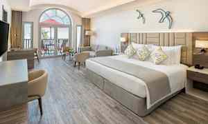 Et moderne hotelværelse med en bred seng, sofa og skrivebord. Store vinduer og balkon i baggrunden. (AI-genereret tekst)