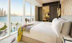 Et lyst hotelværelse med en dobbeltseng, et åbent badeværelse og store vinduer med udsigt over havet. (AI-genereret tekst)