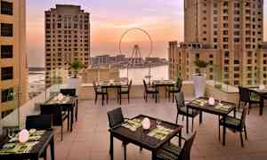 Terrasse med opstillet borde og stole, omgivet af høje bygninger med udsigt til et pariserhjul og havet ved solnedgang. (AI-genereret tekst)