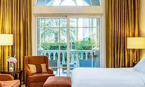 Et hotelværelse med store vinduer, der vender ud mod en terrasse med udsigt til grønne omgivelser. Værelset har en seng, lænestole og guldfarvede gardiner. (AI-genereret tekst)