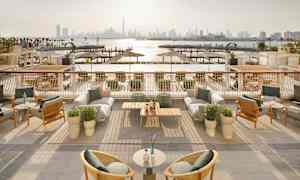 En udendørsterrasse med møbler og udsigt over en havn og byens skyline i baggrunden. (AI-genereret tekst)