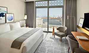 Et moderne indrettet hotelværelse med en stor dobbeltseng og vinduer, der åbner ud til en balkon med udsigt over en byens skyline og vand. (AI-genereret tekst)