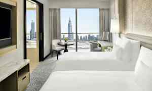 Hotelværelse med to hvide senge, skrivebord og vindue med udsigt over en by med højhuse. (AI-genereret tekst)