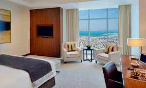 Et moderne hotelværelse med en seng, lænestole, et bord og et arbejdsområde. Store vinduer viser udsigt over by og hav. (AI-genereret tekst)