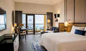 Et hotelværelse med en stor seng, siddeområde og en spiseplads. Store vinduer fører ud til en balkon med udsigt over havet og et stort pariserhjul. (AI-genereret tekst)