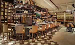 Billede af interiøret i en moderne bar med højryggede stole ved en stor bardisk omgivet af hylder fyldt med flasker og tønder. (AI-genereret tekst)