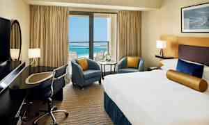 Hotelværelse med en stor seng, skrivebord og udsigt over en blå hav gennem en balkon med glasdøre. (AI-genereret tekst)