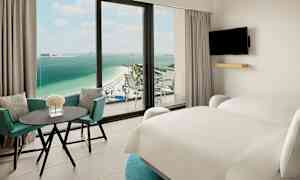 Hotelværelse med to senge, bord og stole samt udsigt over en strand og hav. (AI-genereret tekst)