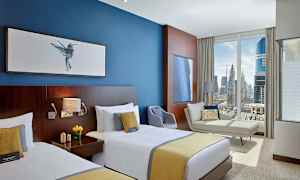 Et hotelværelse med to enkeltsenge, en blå væg med billeder, en sofa, en lænestol og et stort vindue med udsigt over en by. (AI-genereret tekst)