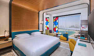 Et hotelværelse med en stor seng, farverige glasmontre og udsigt over strand og by. (AI-genereret tekst)