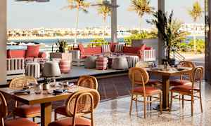 En udendørs restaurant med borde og stole, sofaer og udsigt over vand og huse. (AI-genereret tekst)