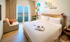 Et hotelværelse med en stor seng, siddeområde og åbent vindue med udsigt over havet. (AI-genereret tekst)