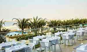 En udendørs restaurant med dækkede borde, med udsigt over palmer, en strand og en bysilhuet i baggrunden. (AI-genereret tekst)