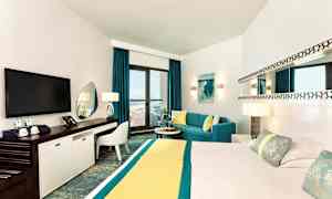 Et moderne hotelværelse med en stor seng, tv, arbejdsområde og en siddegruppe ved et vindue med adgang til en balkon med havudsigt. (AI-genereret tekst)