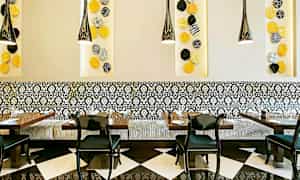 En restaurantinteriør med sort-hvide gulvfliser, mønstrede vægpaneler og en dekorativ siddebænk med sorte stole omkring bordene. Farverige vægdekorationer kan ses på væggen. (AI-genereret tekst)