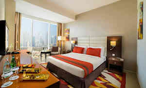 Hotelværelse med stor seng, orange sengetæppe, vindue med udsigt over byen og siddegruppe. (AI-genereret tekst)