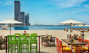 Stole og borde på en strand med udsigt over havet og byens skyline i det fjerne. (AI-genereret tekst)