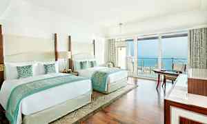 Hotelværelse med to store senge, lyse møbler og havudsigt fra en balkon. (AI-genereret tekst)