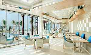 En lys og moderne restaurant med blå puder på stolene og udsigt til en pool og palmer. (AI-genereret tekst)