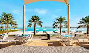 Udendørs serveringsområde med borde, stole og udsigt over tropiske palmer, hvid sandstrand og hav. (AI-genereret tekst)