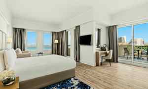 Et moderne hotelværelse med en stor seng, et skrivebord, balkon og udsigt over en blå himmel og bygninger. (AI-genereret tekst)