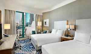 Et hotelværelse med to dobbeltsenge, et skrivebord med stol og en balkon med udsigt over moderne bygninger. (AI-genereret tekst)