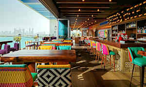 En moderne restaurant med farverige stole og barstole, træmøbler og udsigt over en bysilhuet ved vandet. (AI-genereret tekst)